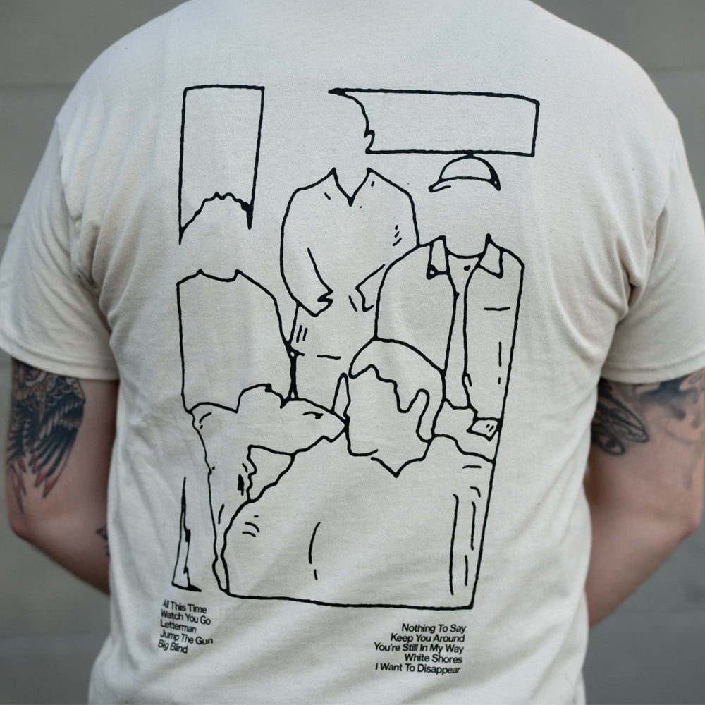 A person wearing a beige The Story So Far Gildan Heavy T-shirt featuring a black line drawing of five abstract, faceless figures and small printed text near the bottom; the wearer’s tattooed arms are visible.