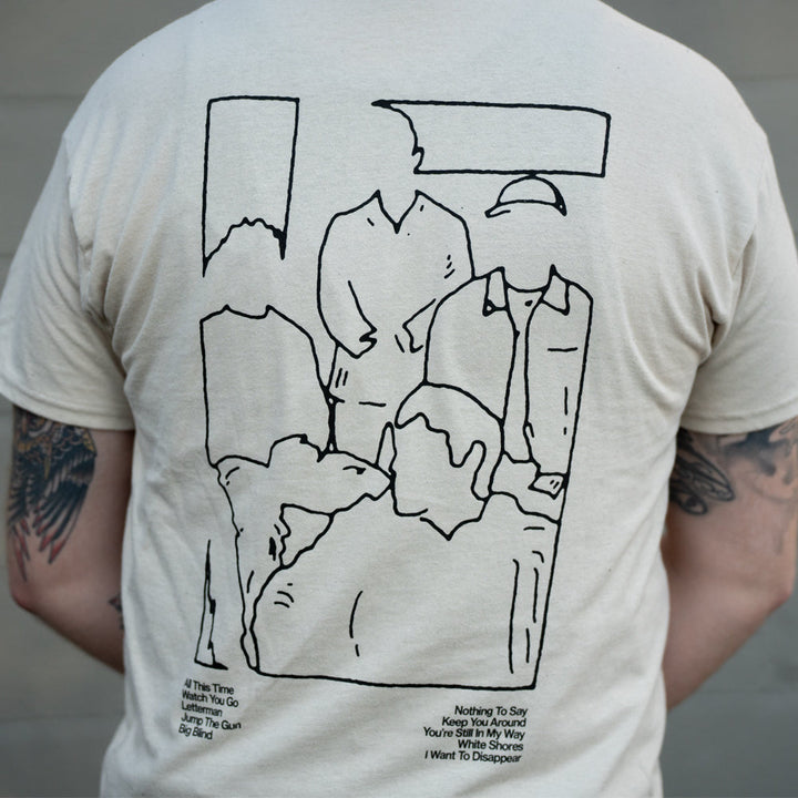 A person wearing a beige The Story So Far Gildan Heavy T-shirt featuring a black line drawing of five abstract, faceless figures and small printed text near the bottom; the wearer’s tattooed arms are visible.