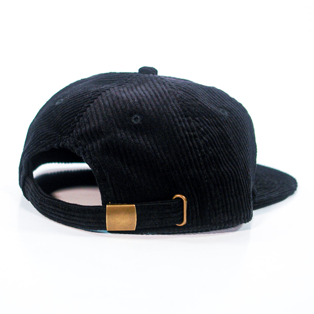 A black Spanish Love Songs Class Chord Cap is shown from the back, featuring an adjustable gold-colored metal clasp. This unstructured six-panel cap is made from 100% cotton corduroy and set against a white background.