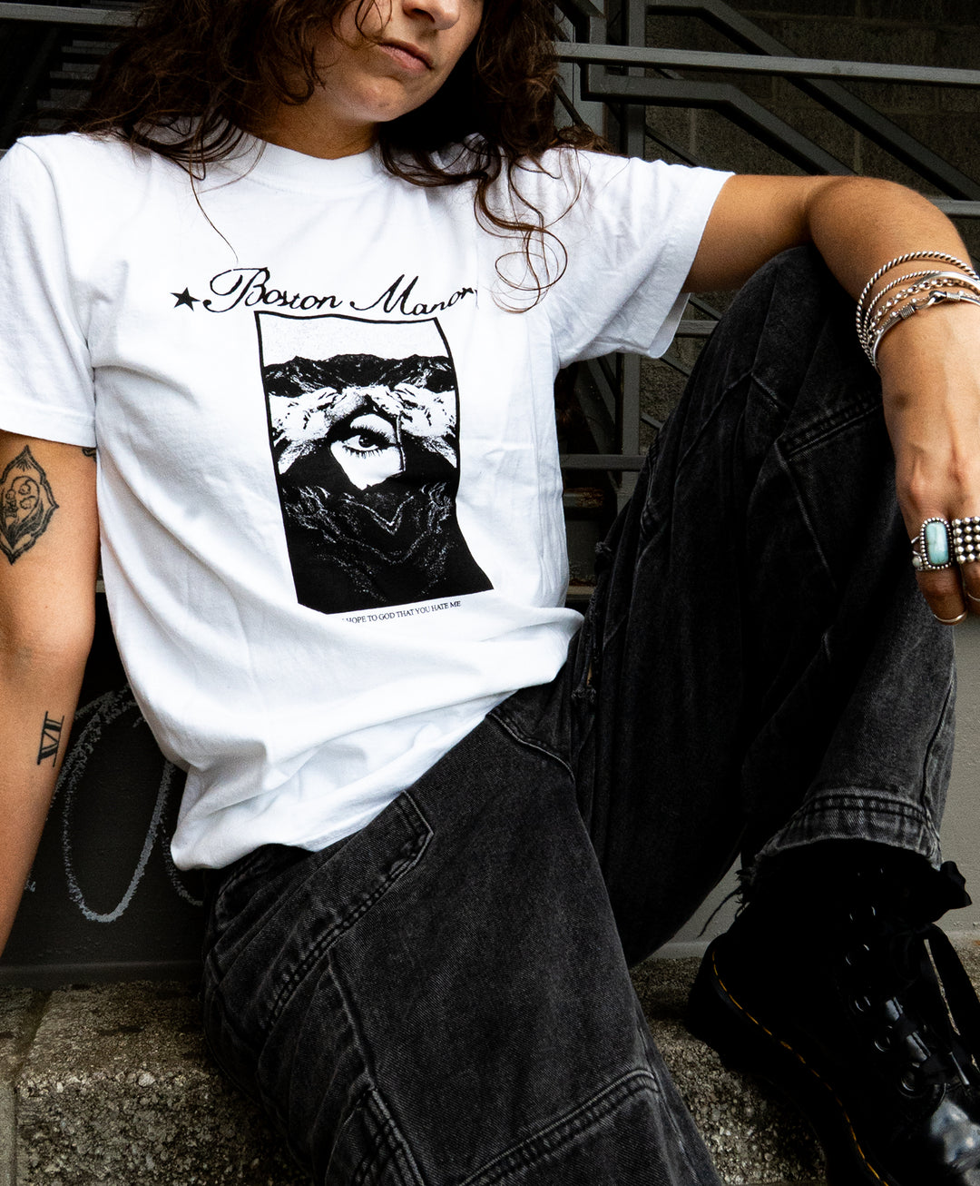 Person sitting on concrete steps, wearing a white Comfort Colors printed t-shirt that says Bison Maron with an illustrated face, black jeans, boots, and silver jewelry. The background includes metal railings and a brick wall.