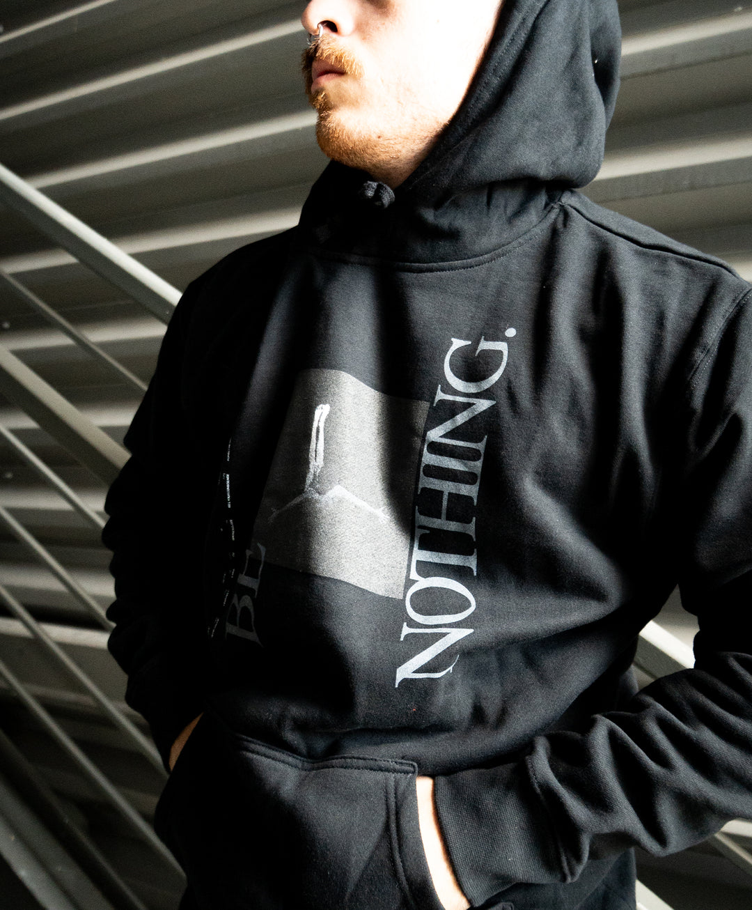A person wearing a black hoodie from Independent Trading Company, with the word NOTHING. printed on it, stands with hands in the front pocket under a metal staircase—a stylish example of midweight pullovers and printed pullovers combined.