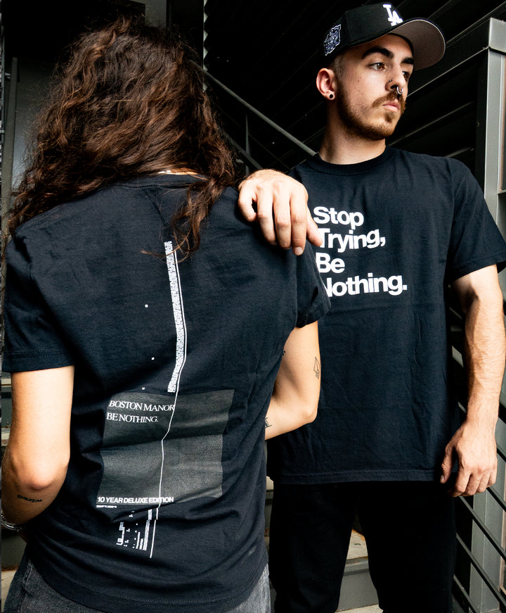 Two people in black Comfort Colors t-shirts stand on stairs. One faces away, showing graphic text on the back; the other faces forward, with Stop Trying, Be Nothing. printed on the shirt. Both have dark hair; one wears a black cap.