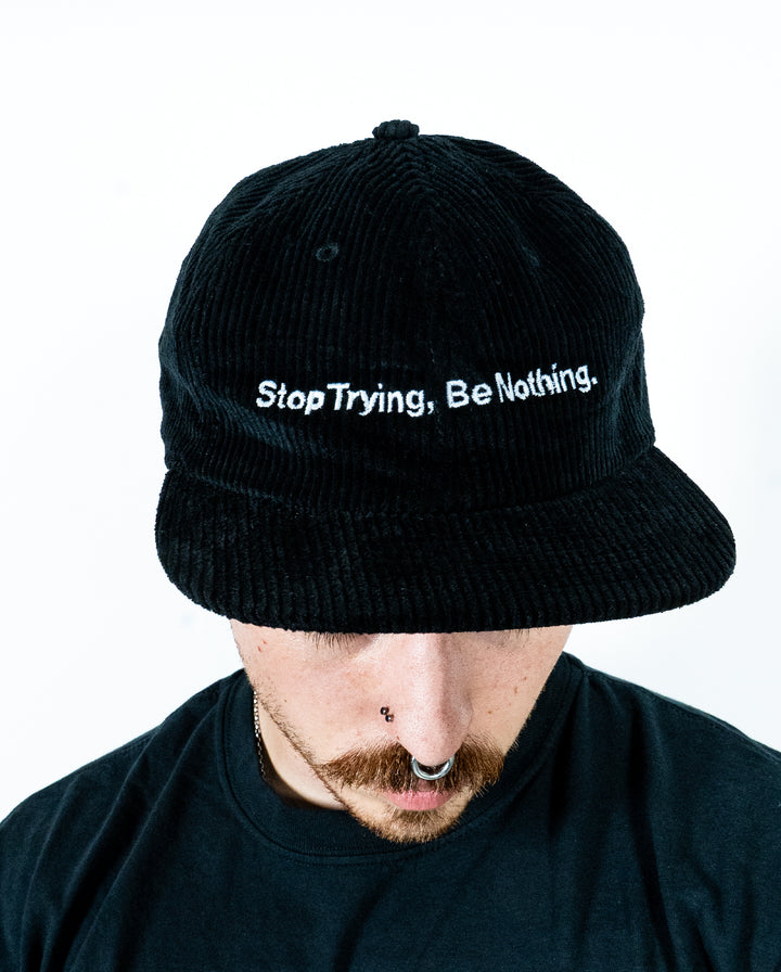 A person with facial piercings and a mustache looks down, wearing a black corduroy hat from Class Caps that says Stop Trying, Be Nothing. The background is plain white.