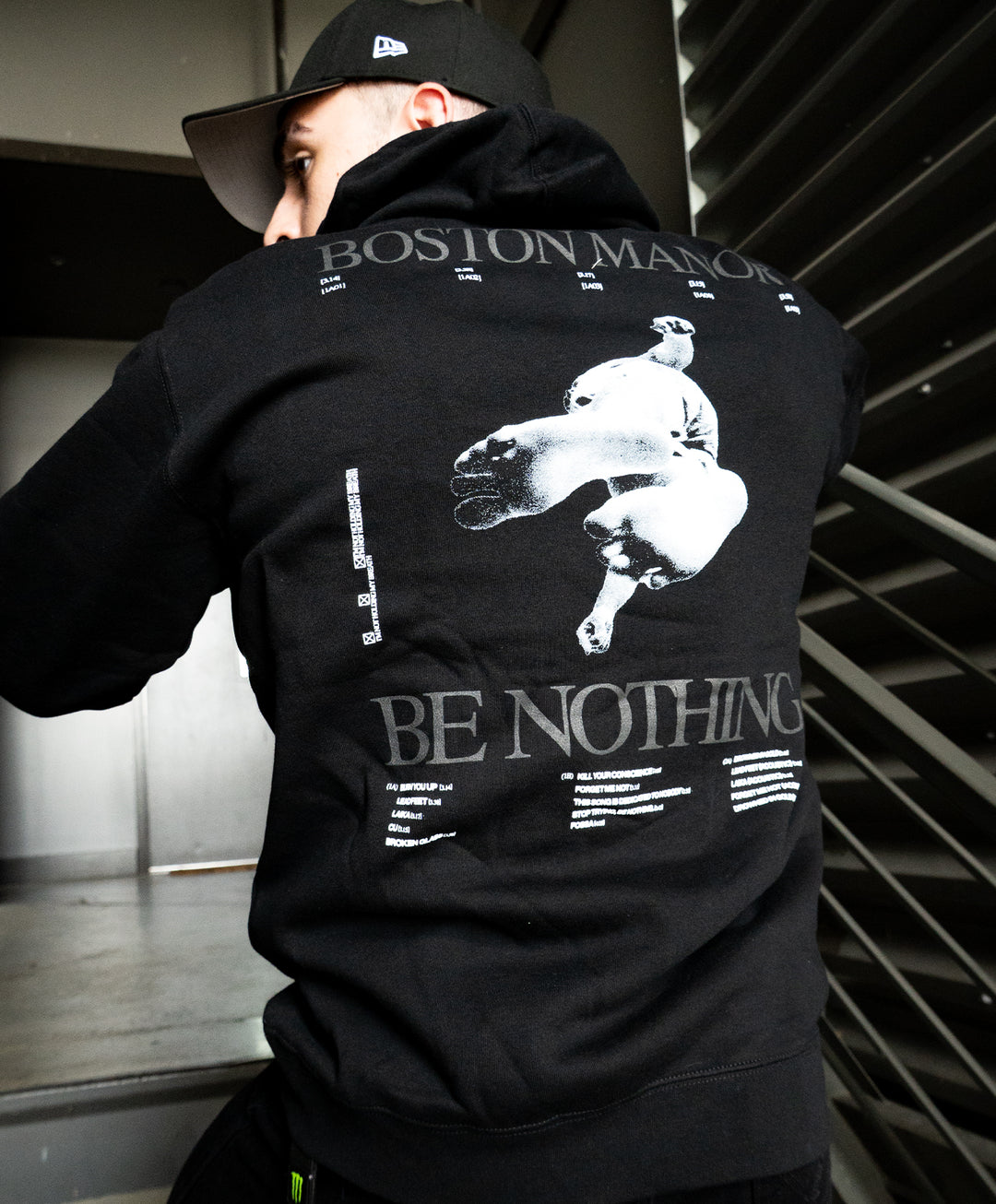 A person wearing a black midweight pullover hoodie from Independent Trading Company, featuring Boston Manor and Be Nothing printed on the back along with graphic and text designs, stands near a metal staircase.