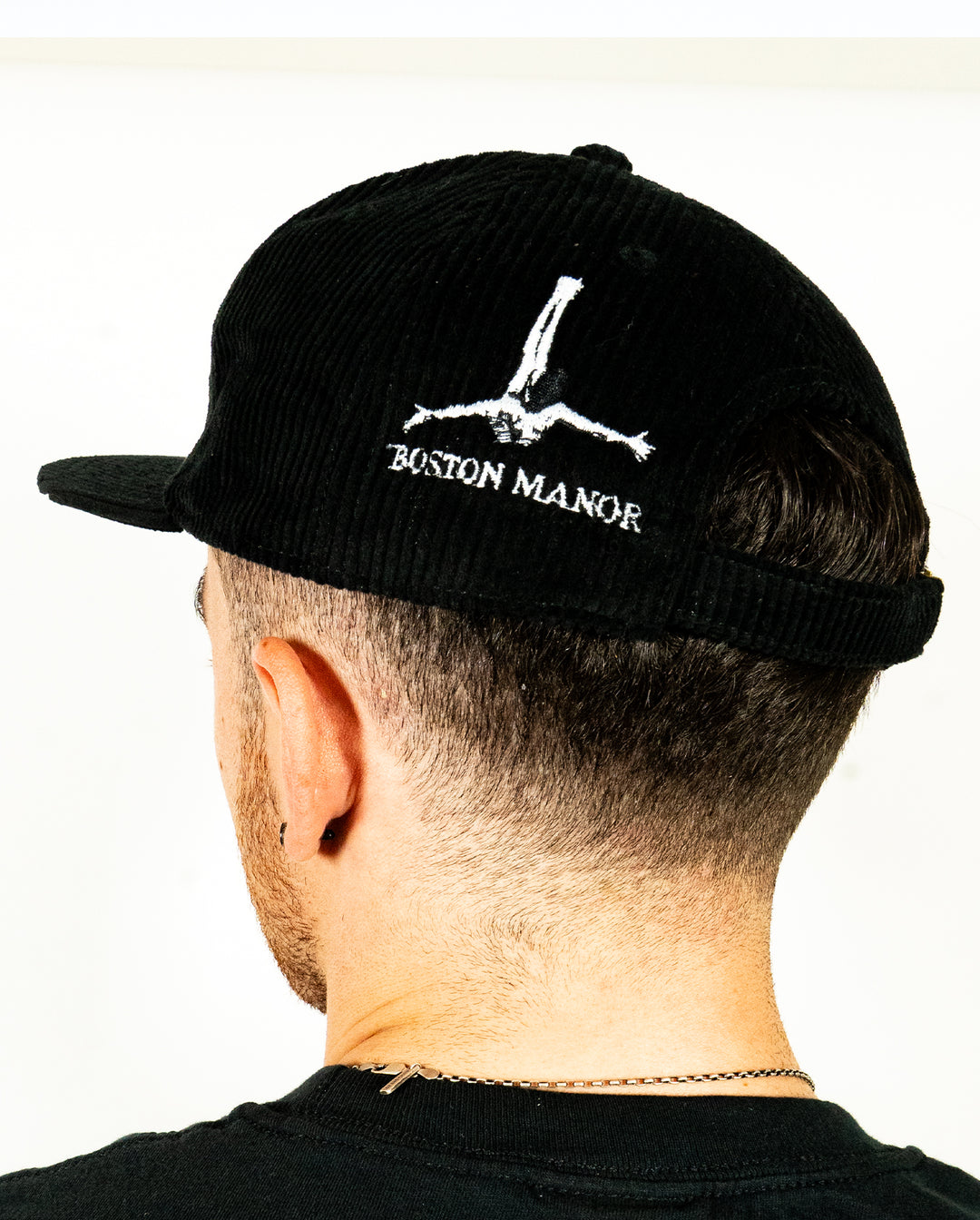 A person wearing a black corduroy cap from Class Caps with Boston Manor and an upside-down figure embroidered on the back, shown from behind.