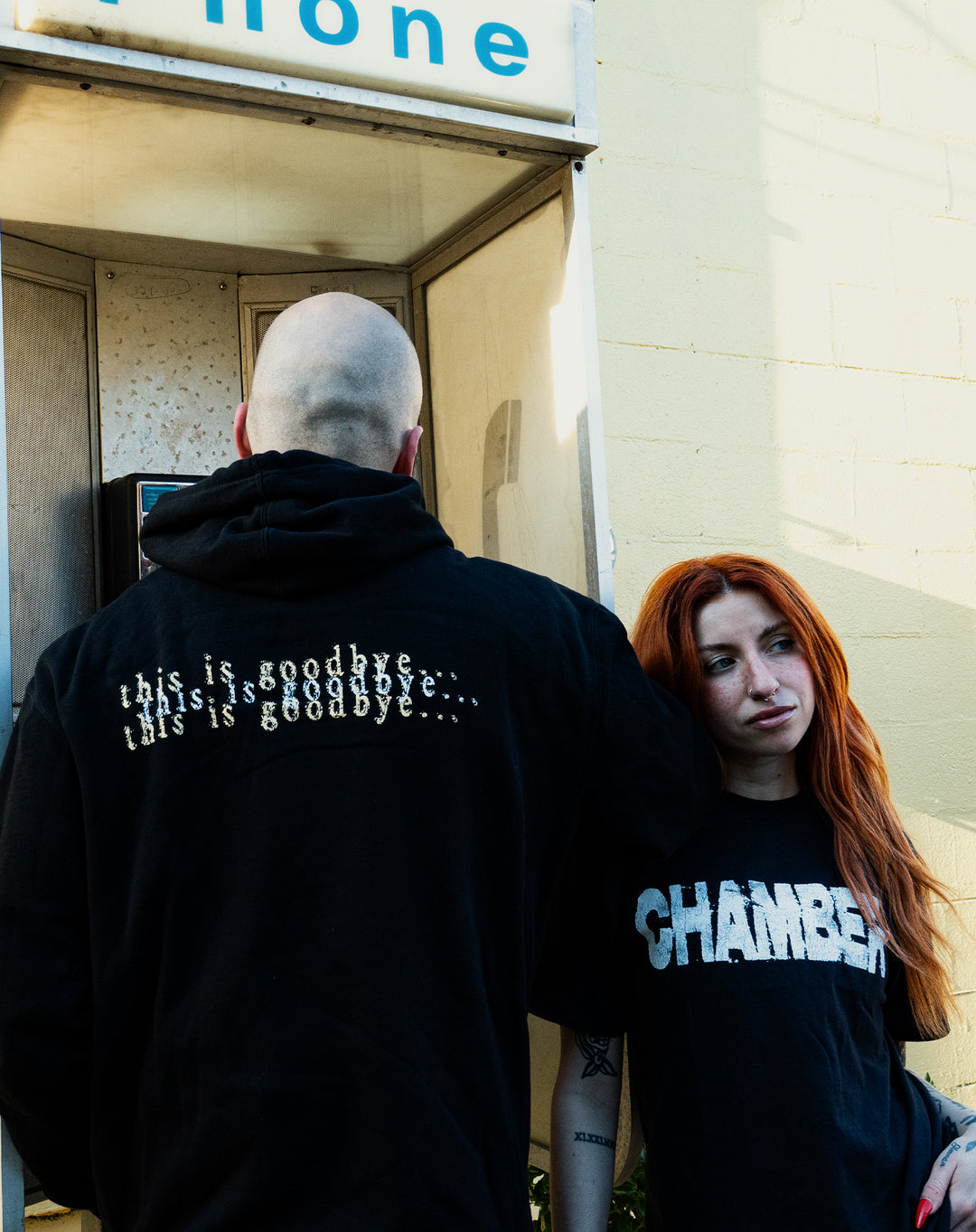 A bald person in a black hoodie with "this is goodbye..." on the back faces a phone booth. Nearby, a red-haired person in a Chamber Comfort Colors printed t-shirt that says "CHAMBER" leans against the wall, looking away.