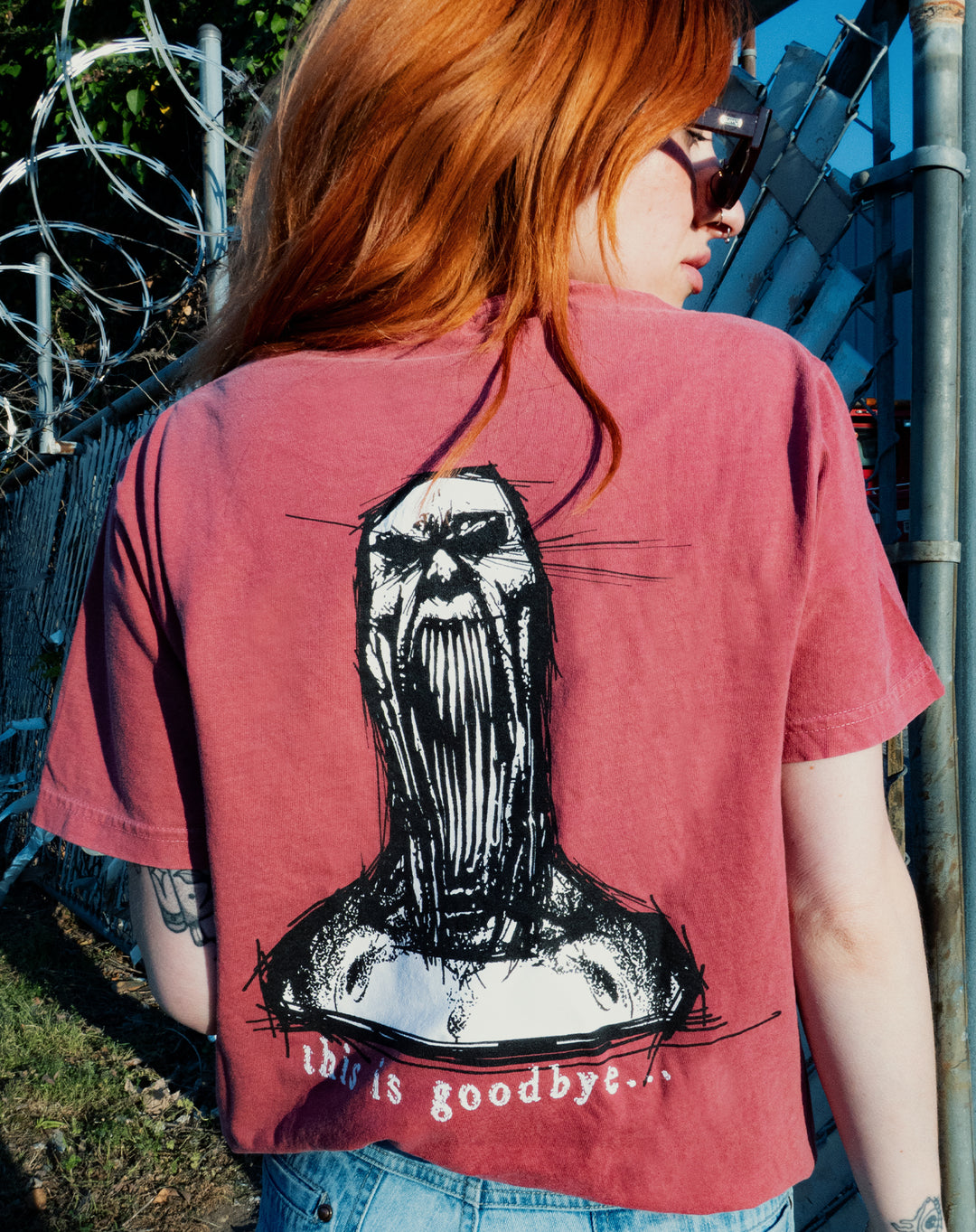 A person with long red hair and sunglasses stands by a chain-link fence, wearing a pink Chamber Comfort Colors tee with a graphic of a screaming figure and “this is goodbye…” printed on the back.