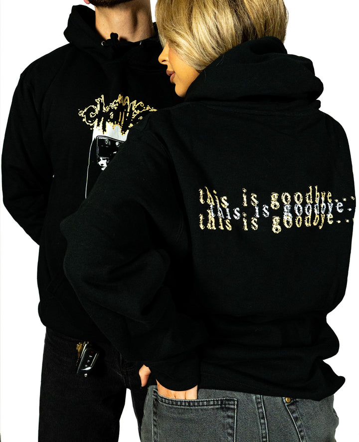 Two people in black Chamber custom apparel: the woman’s printed sweatshirt reads “this is goodbye...” in a gold, glitched font, while the man wears an abstract graphic. Both sport Chamber hoodies and face away from the camera.