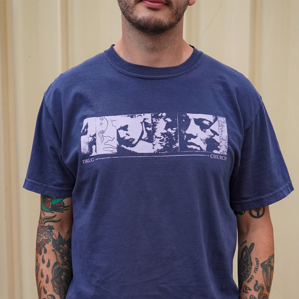 A tattooed person wears a blue Drug Church Comfort Colors T-shirt with three abstract, monochrome faces in rectangles and DRUG CHURCH below, pictured against a beige wall.