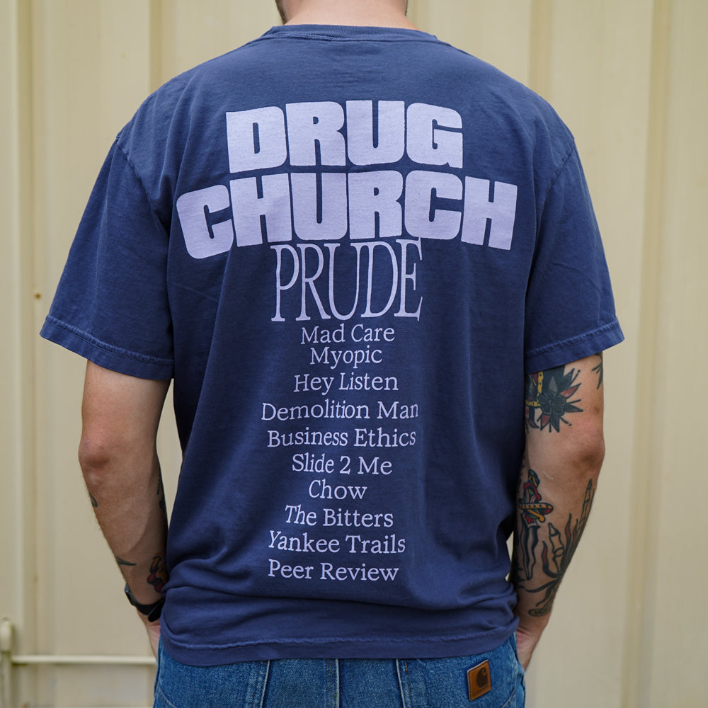 A person with visible tattoos stands before a beige wall, wearing a navy blue Comfort Colors t-shirt by Drug Church featuring DRUG CHURCH PRUDE and song titles printed on the back.