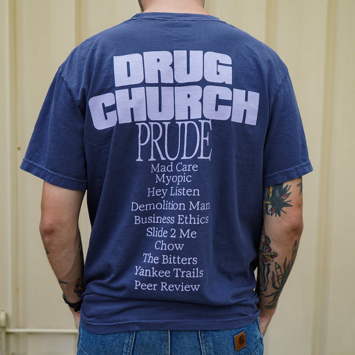 A person with visible tattoos stands before a beige wall, wearing a navy blue Comfort Colors t-shirt by Drug Church featuring DRUG CHURCH PRUDE and song titles printed on the back.