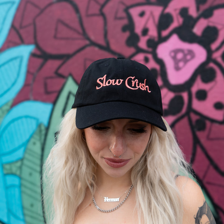 A person with long blonde hair and facial piercings wears a black Slow Crush Logo Dad Hat and a Herman necklace, standing in front of a colorful mural with floral and leaf patterns.