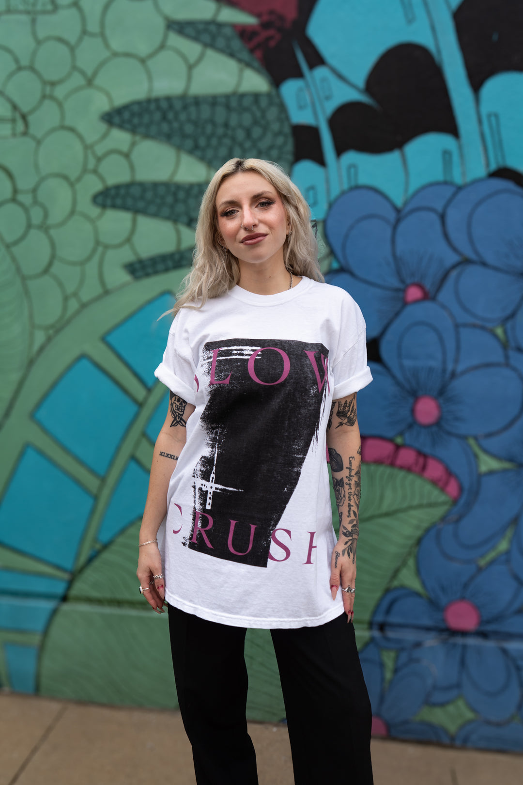 A woman with blonde hair and tattoos poses confidently before a vibrant mural, wearing a Slow Crush Comfort Colors white shirt featuring a bold black and pink graphic design, paired with black pants.