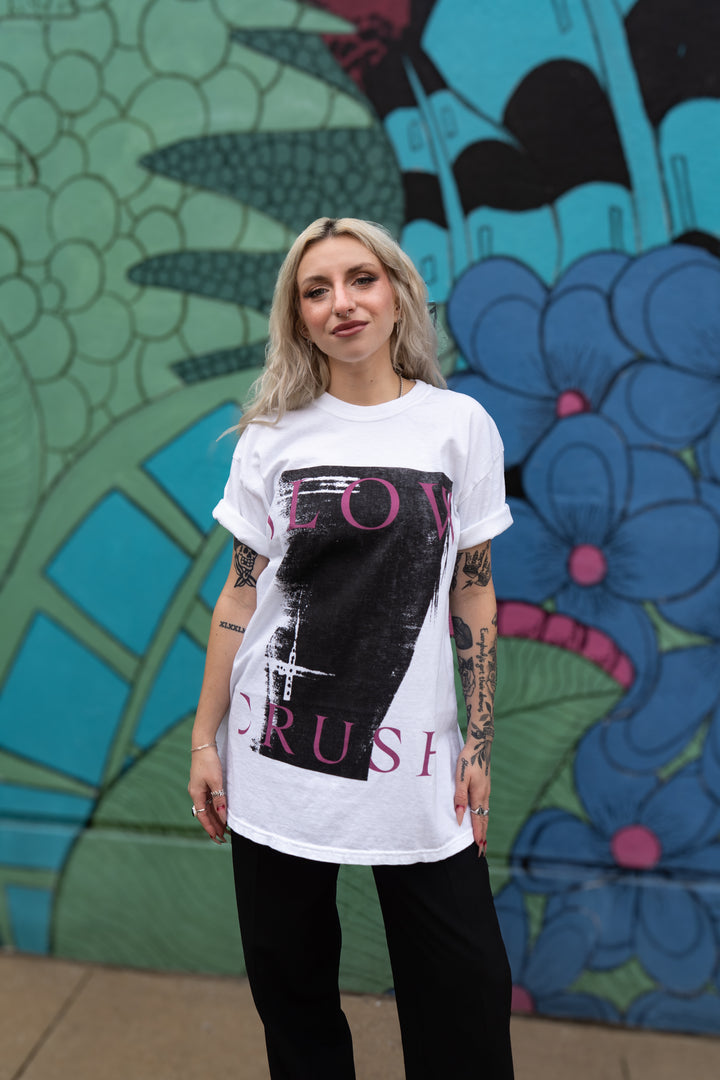 A woman with blonde hair and tattoos poses confidently before a vibrant mural, wearing a Slow Crush Comfort Colors white shirt featuring a bold black and pink graphic design, paired with black pants.