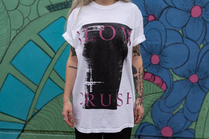 A tattooed individual wears a white Slow Crush shirt with pink and black text, standing before a vibrant mural featuring blue flowers and green geometric patterns.