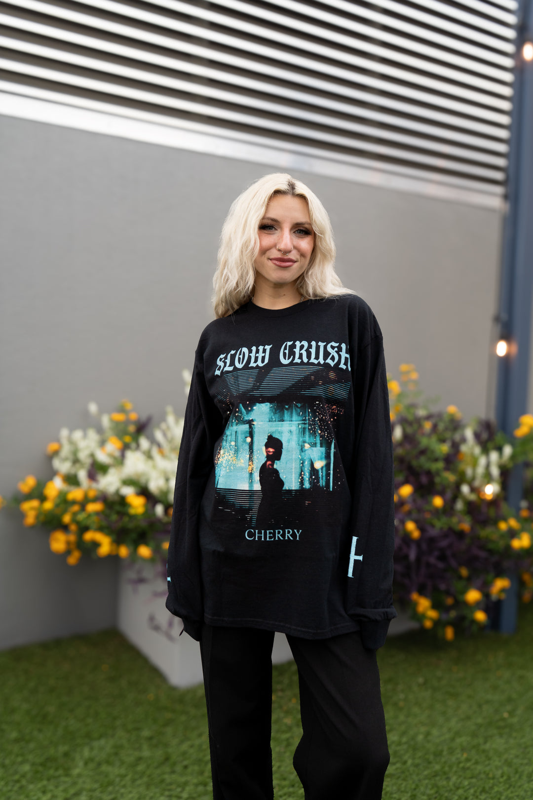 A person with shoulder-length blond hair stands on grass, wearing a black Slow Crush Cherry print t-shirt and black pants. Planters of yellow and white flowers are visible behind them.