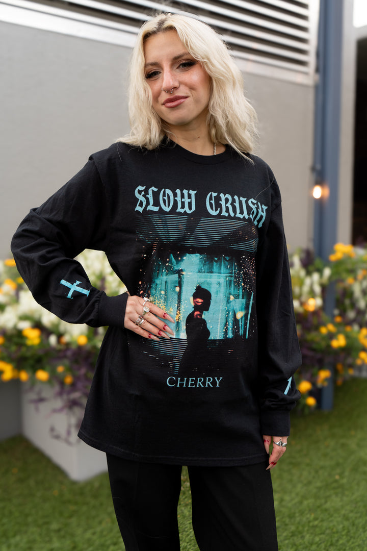 A blonde woman with a nose ring smiles outdoors on grass, wearing a black Slow Crush Cherry Gildan t-shirt. Yellow and white flowers are visible in the background.