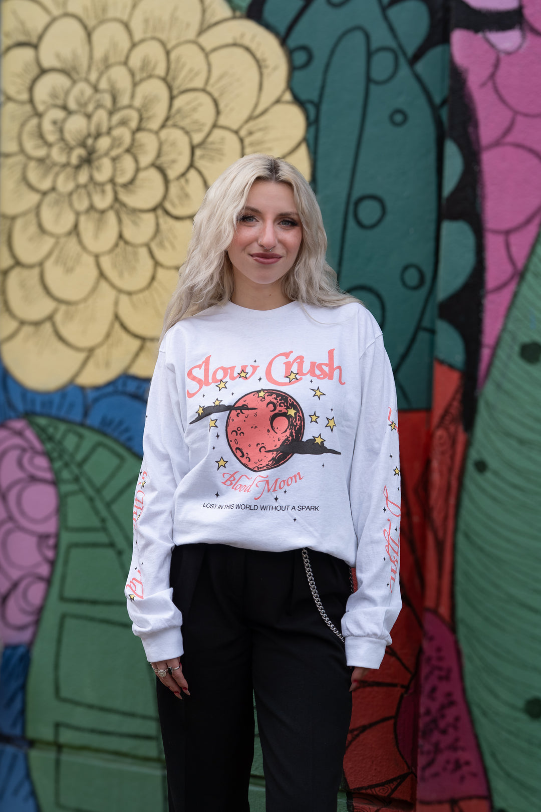 A young woman with long blonde hair stands before a colorful mural of large flowers and abstract shapes, wearing a white Slow Crush printed shirt with red graphics and black pants, smiling softly at the camera.