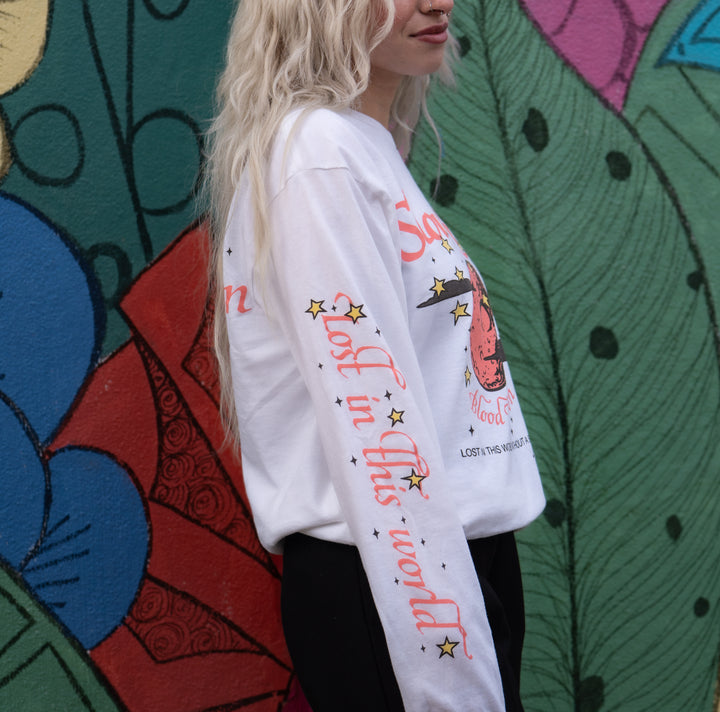 A woman with long, wavy blond hair wears a white Slow Crush printed shirt featuring decorative text and graphics. The shirt sleeve reads lost in this world with stars, as she stands before a colorful, patterned mural.