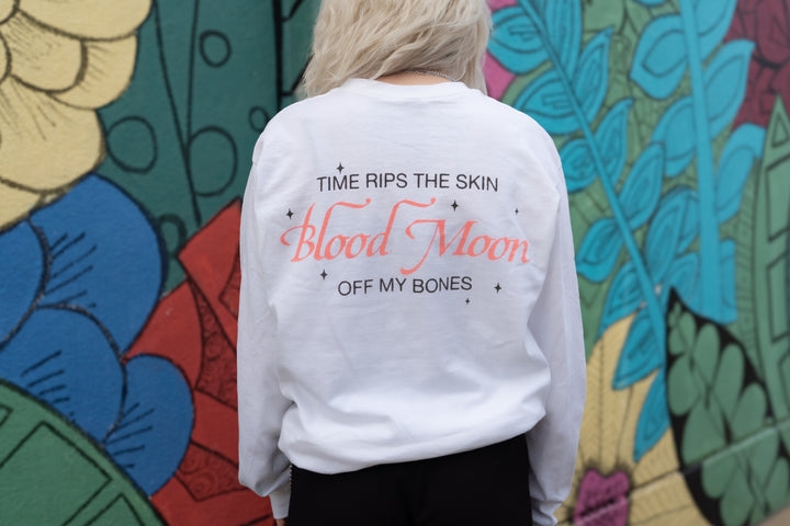A person with light blonde hair wears a white Slow Crush printed shirt that says TIME RIPS THE SKIN Blood Moon OFF MY BONES, standing in front of a colorful floral mural.