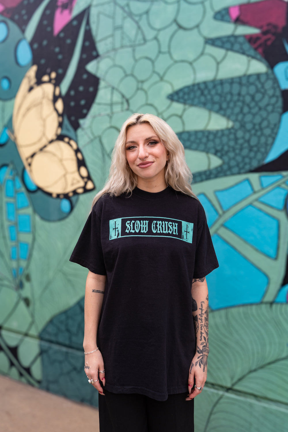 A blonde woman with visible tattoos smiles in front of a vibrant mural with a butterfly and green leaves, wearing a black Slow Crush t-shirt.