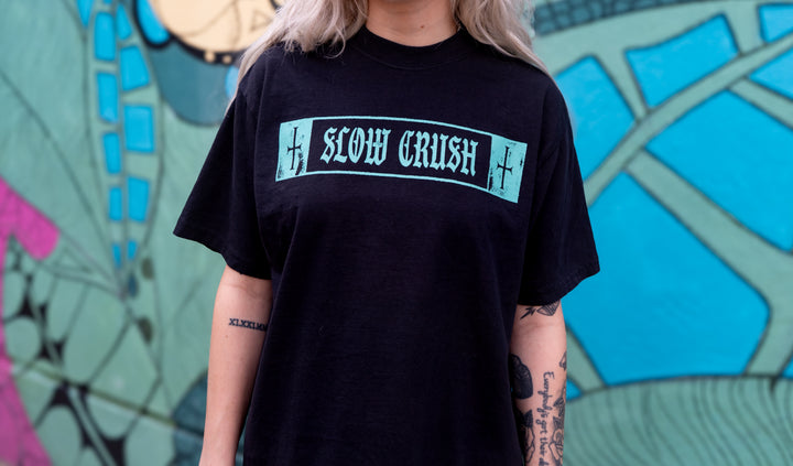 A person wearing a black Slow Crush Comfort Colors printed t-shirt stands before a colorful abstract mural. Their tattooed arms rest at their sides, and only their torso and part of their face are visible in the shot.