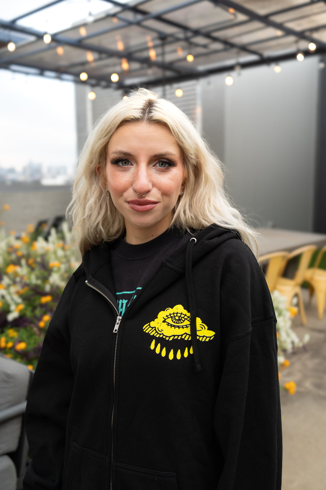 A young woman with long blonde hair stands outdoors under string lights, wearing a Slow Crush Independent Midweight Zip hoodie with a yellow graphic. Flowers and yellow chairs are visible in the background.