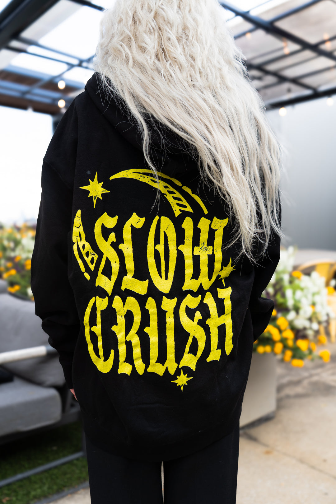 A person with long, wavy platinum blonde hair wears a black Slow Crush hoodie featuring large yellow text and star graphics on the back, standing outdoors near flowers and patio furniture.