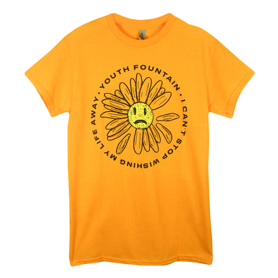 image of a gold tee shirt on a white background. tee has center print of a daisy with a sad face in the center. around the daisy says youth fountain i can't stop wishing my life away