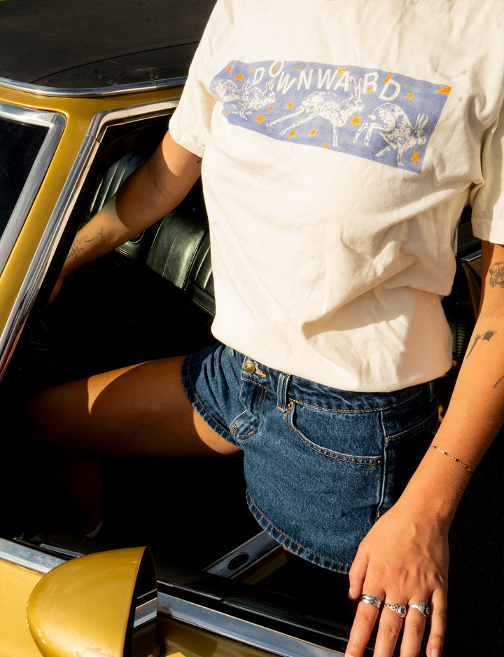 Someone wearing a white DOWNWARD brand printed shirt from Comfort Colors and denim shorts sits in a vintage yellow car, with sunlight accentuating their outfit and jewelry.