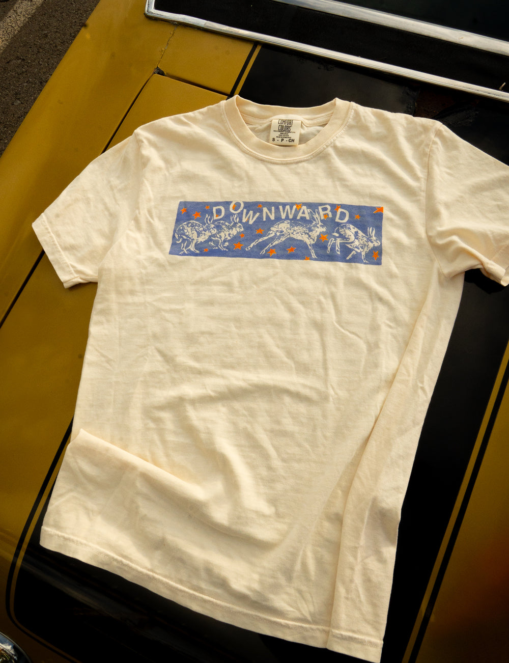 A white Comfort Colors t-shirt by DOWNWARD, featuring a blue "DOWNWARD" graphic and playful dinosaur illustrations, laid flat on the hood of a yellow car.