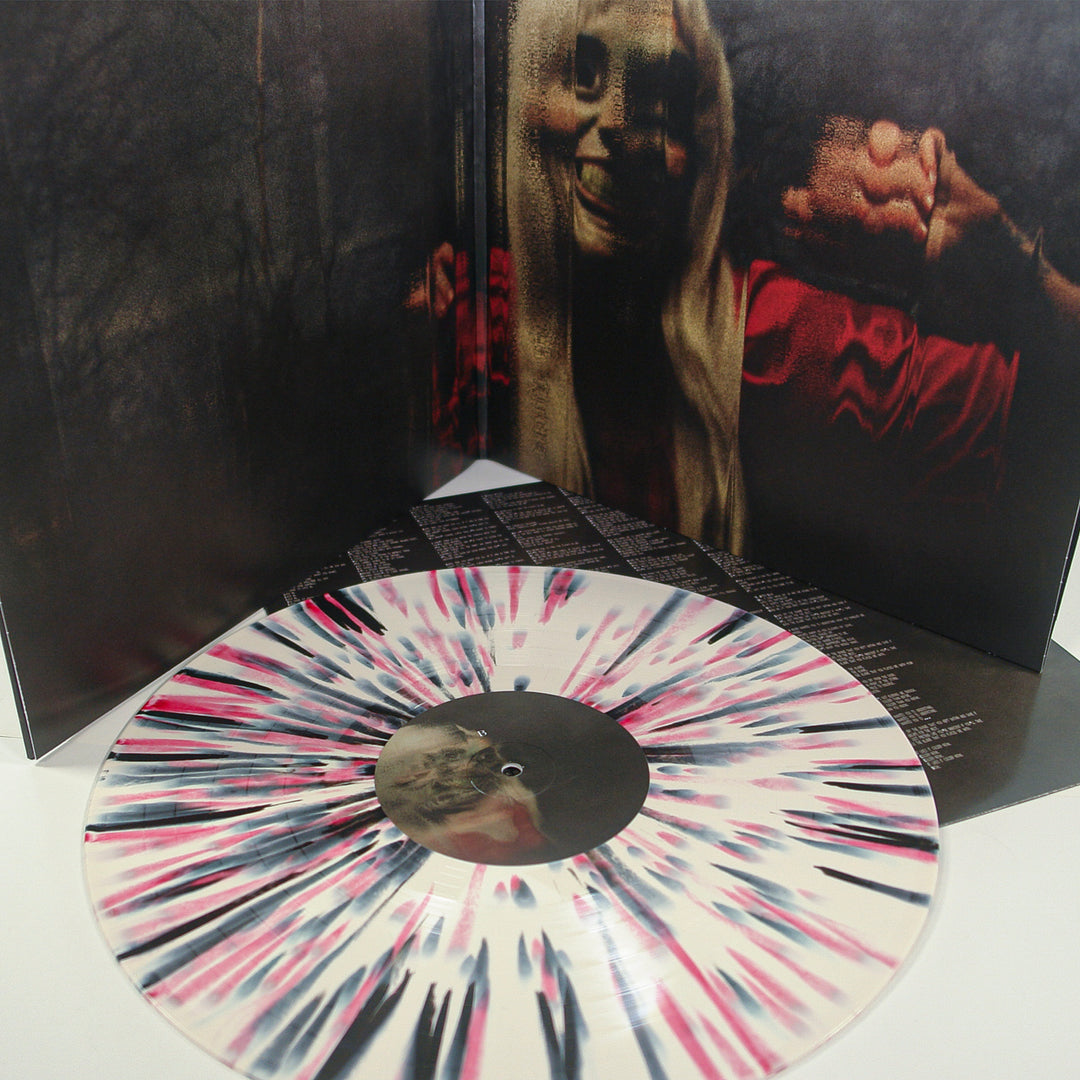 A bone vinyl record with pink and black splatter resting in front of an open gatefold album cover featuring an image of a smiling, long-haired person in a red shirt.