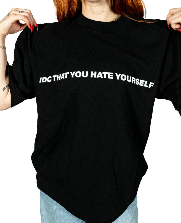 A person wears a black Knocked Loose T-shirt made from premium USA cotton, featuring white text that reads, IDC THAT YOU HATE YOURSELF. They hold the sides of the garment to clearly display the message.