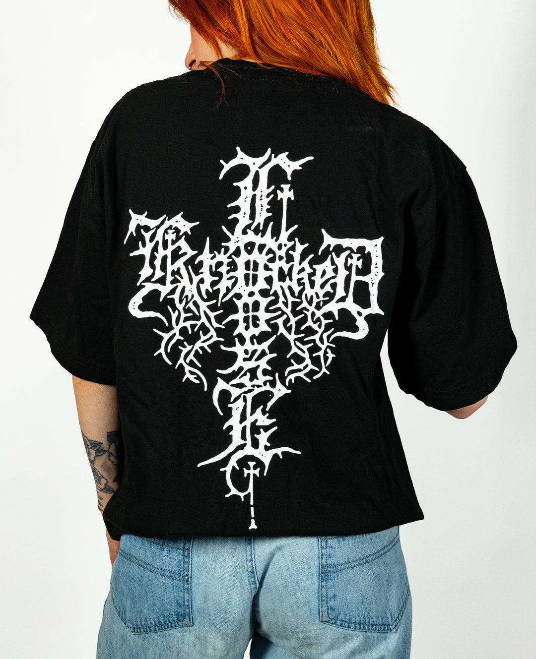 Person with long red hair wears a black Knocked Loose T-shirt made from premium USA cotton, featuring an intricate white design on the back. Paired with light blue jeans, the person is turned away to highlight the graphic on this garment dyed tee.