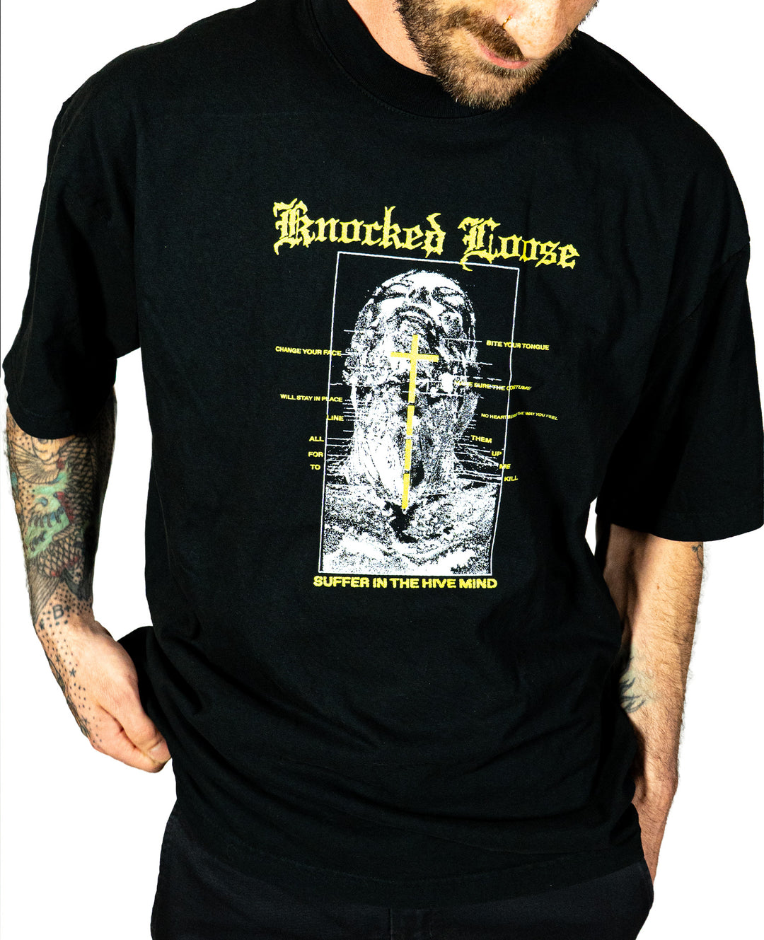 Wearing a black Knocked Loose tee with a distressed face graphic, bold yellow text, and "SUFFER IN THE HIVE MIND," this spacious LA Apparel shirt in premium, shrink-free cotton shows off their tattooed arm.