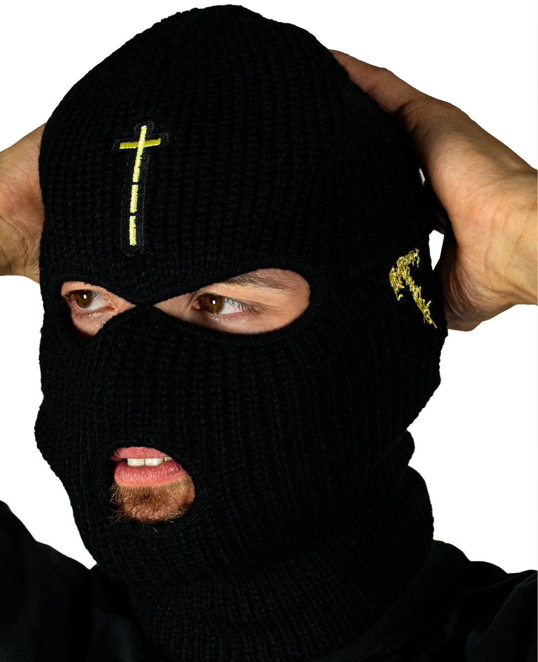 A person wears a black Knocked Loose knit balaclava with a yellow cross and small yellow stitching, holding the sides with both hands. This presale item ships starting 04/24/2026. White background.