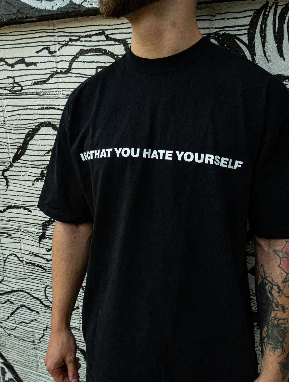 A person stands in front of a mural, wearing a black Knocked Loose T-shirt made from premium USA cotton, with "NOT THAT YOU HATE YOURSELF" in white on the chest. Only the lower half of their face and torso are visible.