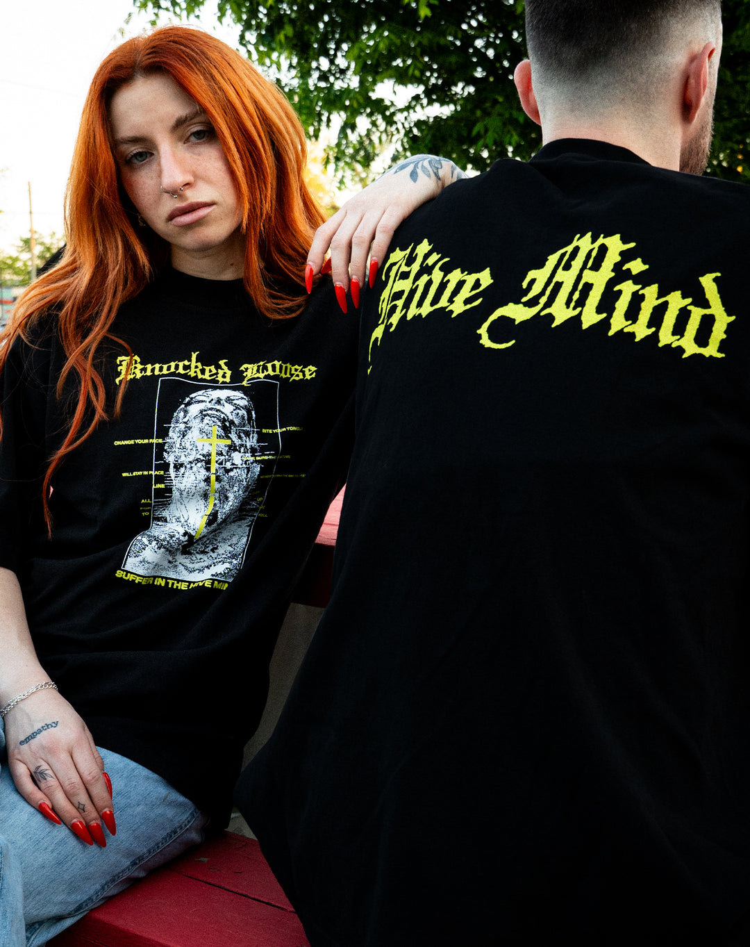 Two people sit outdoors on a red bench. The woman with long red hair and tattoos wears a black shirt with yellow graphics. The man, back turned, wears a Knocked Loose LA Apparel T-Shirt with “Hive Mind” in yellow gothic text. Green trees are behind them.
