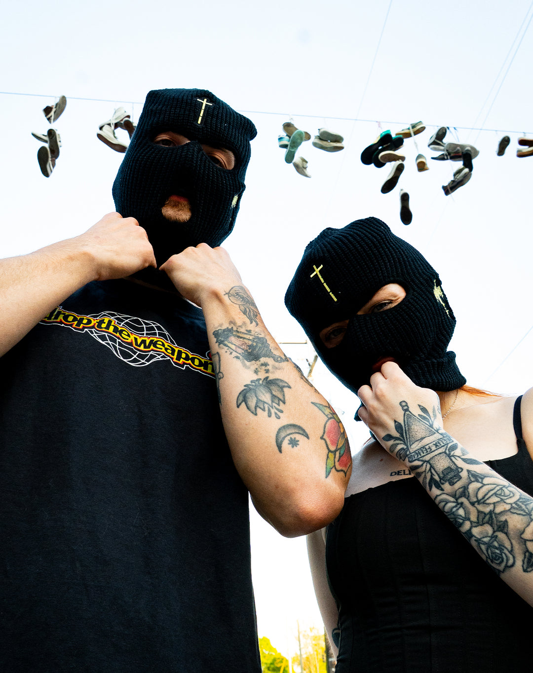 Two tattooed people in black ski masks, one with a cross, stand under shoes hanging from wires. The person on the left wears a black Knocked Loose T-shirt and has a tattooed arm. This presale item ships 04/24/2026.