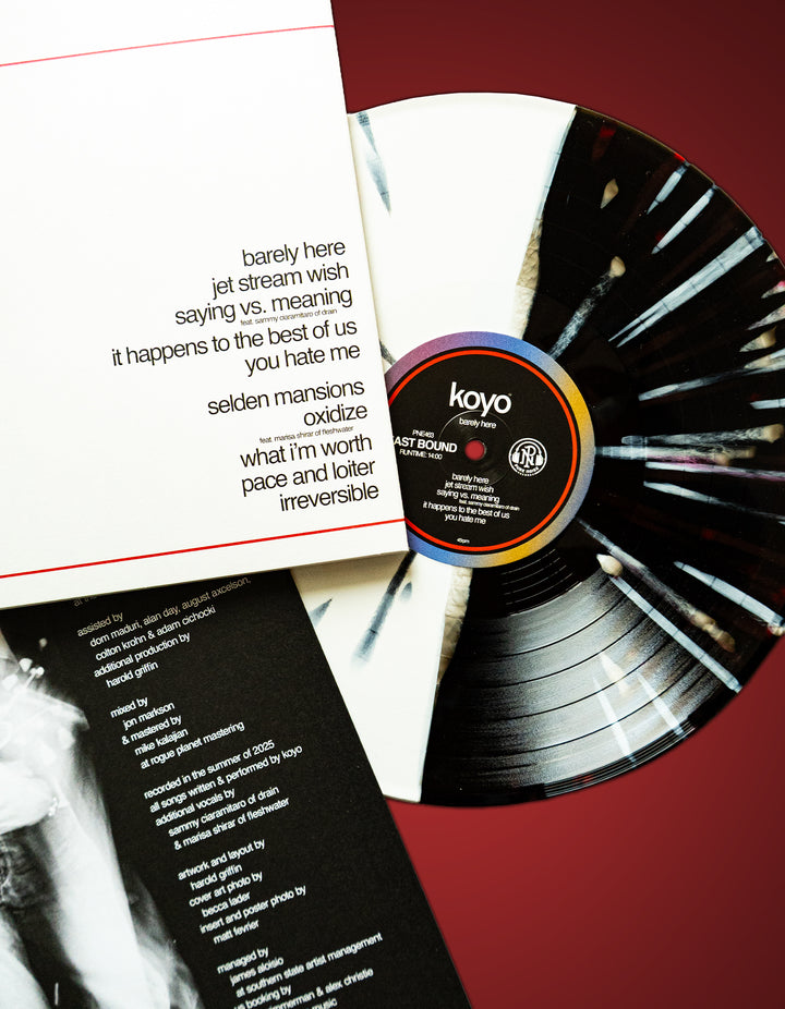 A black and white splatter KOYO Barely Here vinyl LP, partially out of its sleeve, rests on a red surface. The visible tracklist includes songs like “barely here,” “pace,” and “loyer irreversible.”.