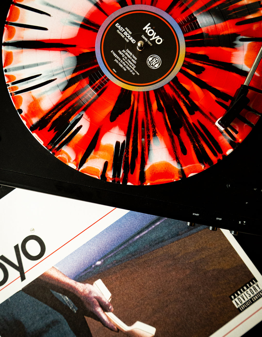 A close-up of a KOYO vinyl LP in red, black, and white spinning on a turntable. Below is the KOYO album cover featuring a hand holding white objects and a parental advisory label.