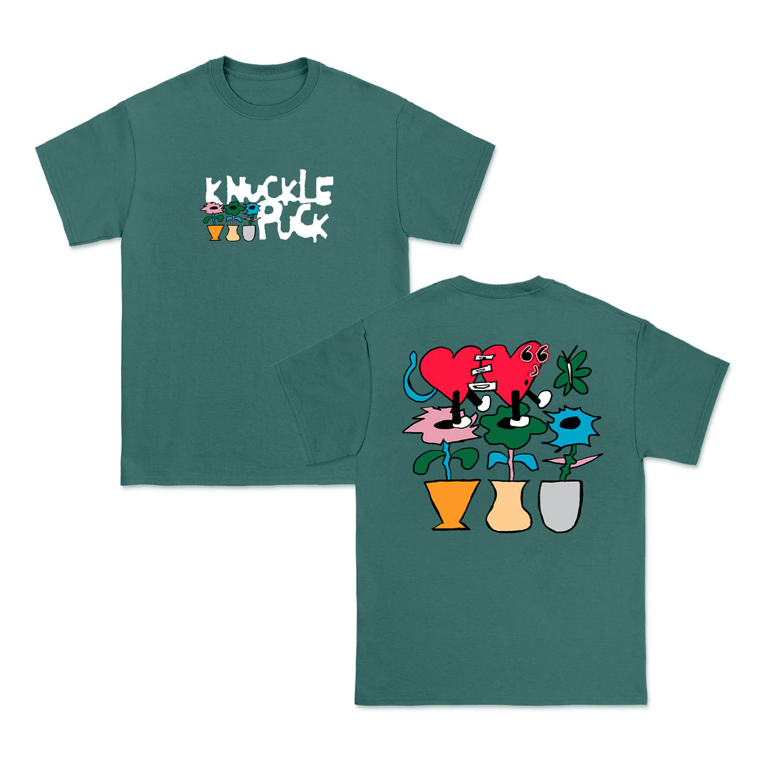 Front and back of a teal Knuckle Puck Comfort Colors shirt. The front has KNUCKLE PUCK in white with small cartoon flowers; the back shows four colorful flowerpots with whimsical flowers and a red heart character.