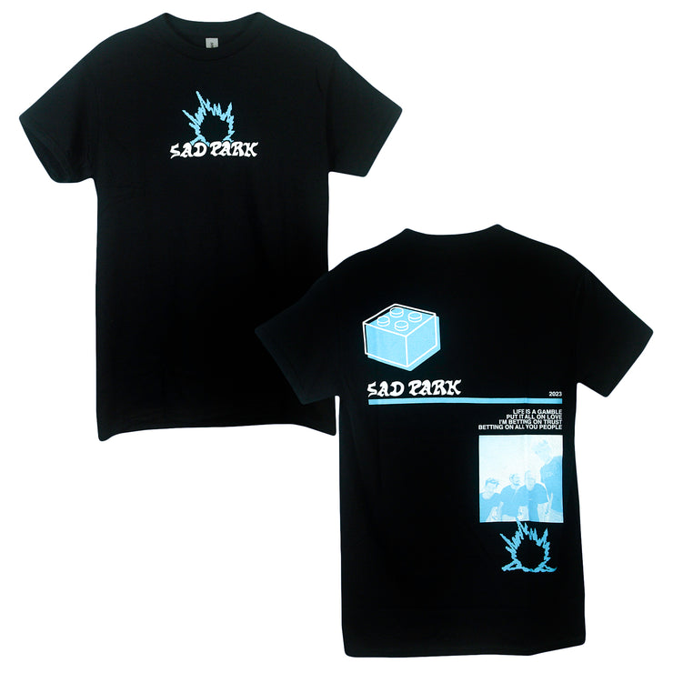 image of the front and back of a black tee shirt on a white background. front is on the left and has a small center chest print that says sad park, with a blue flame above it. the back is on the right and has a full print of a lego piece, sad park, a blue and a white photo of the band