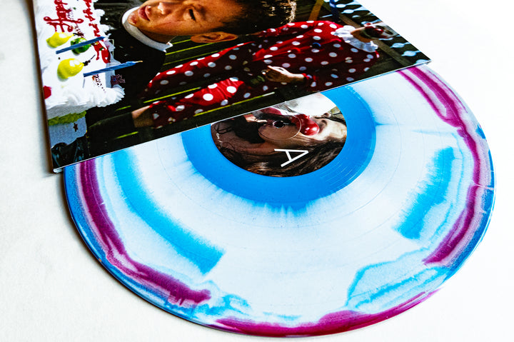 A colorful Glitter Vinyl LP by Drug Church with blue and pink swirls, partially covered by an album cover showing a child blowing out candles on a birthday cake.