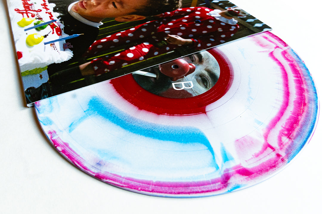 A vibrant America Marble vinyl record by Drug Church, featuring blue, pink, and white swirls, is partially slipped from its sleeve that displays a child before a birthday cake with a clown.