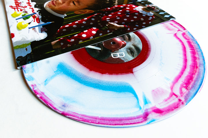 A vibrant America Marble vinyl record by Drug Church, featuring blue, pink, and white swirls, is partially slipped from its sleeve that displays a child before a birthday cake with a clown.