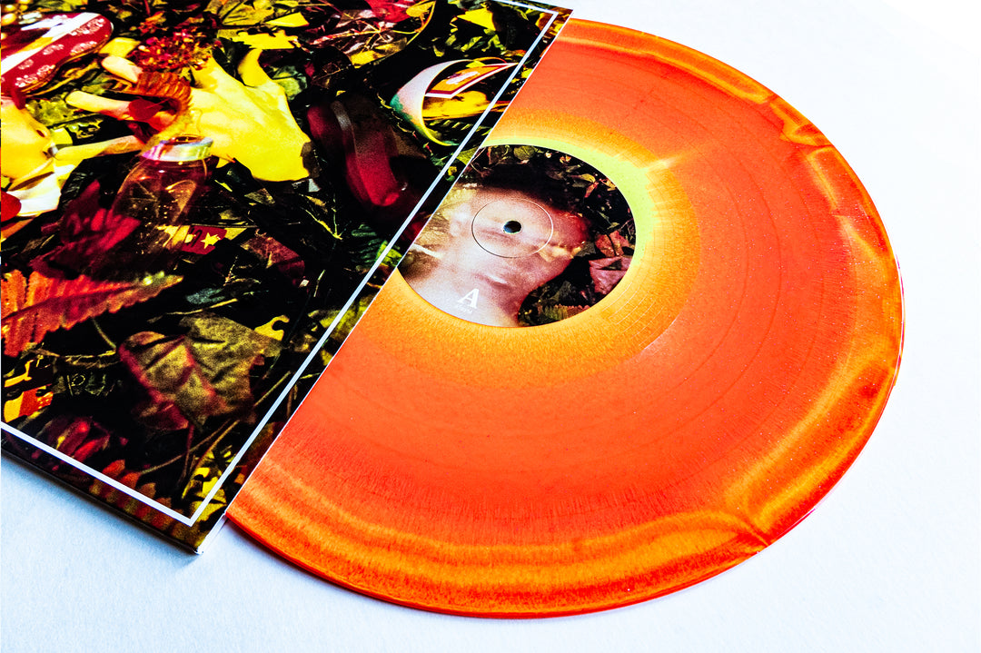 An orange and yellow Glitter Vinyl LP by Drug Church is partially slid out of a colorful, abstract album cover, with a close-up face on the label and a bright white background—ideal for your next album presale.