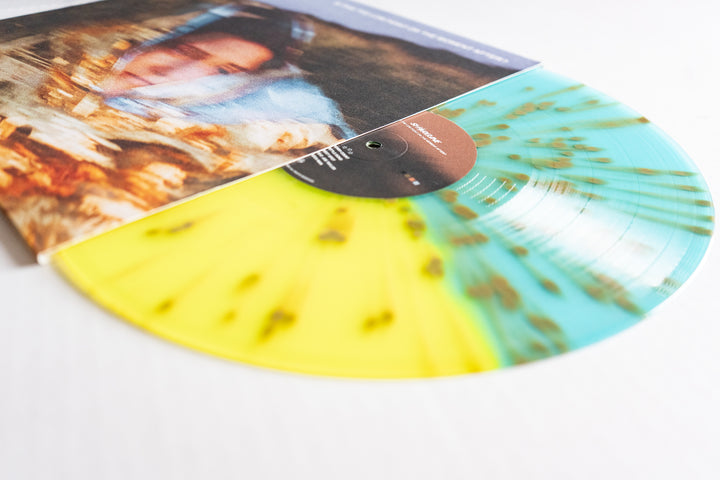 A vibrant Shoreline Gold Splatter Vinyl with yellow, teal, and brown is partially pulled from its album cover, which displays a blurred image of a person’s face.