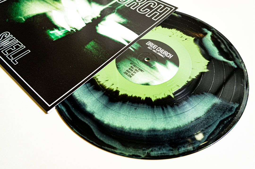 A Drug Church Swell Glitter Vinyl LP in mint/black/olive with a black and green splatter design is partially out of its album cover, which features abstract green and black artwork with "DRUG CHURCH" and "SWELL" text.