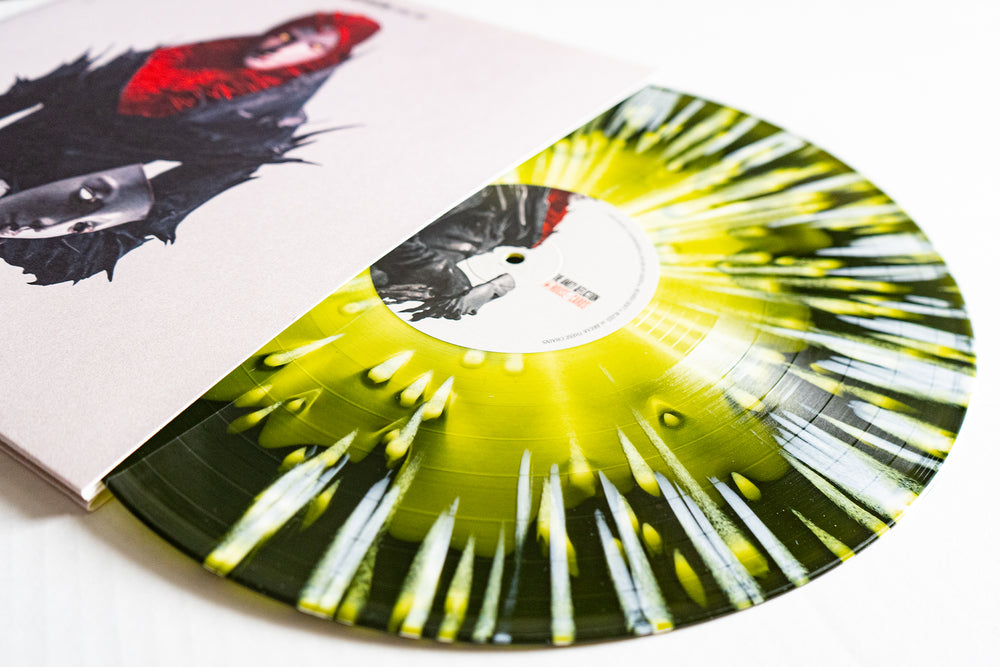 The Amity Affliction’s Yellow in Green with White Splatter vinyl LP is available for presale, featuring a moody cover with two figures in dark feathered clothing and a splatter-patterned House Of Cards record partly out of its sleeve.